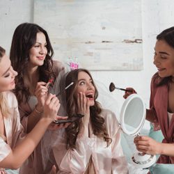 Multiethnic women putting makeup with cosmetic brushes on excited and happy bride at bachelorette party